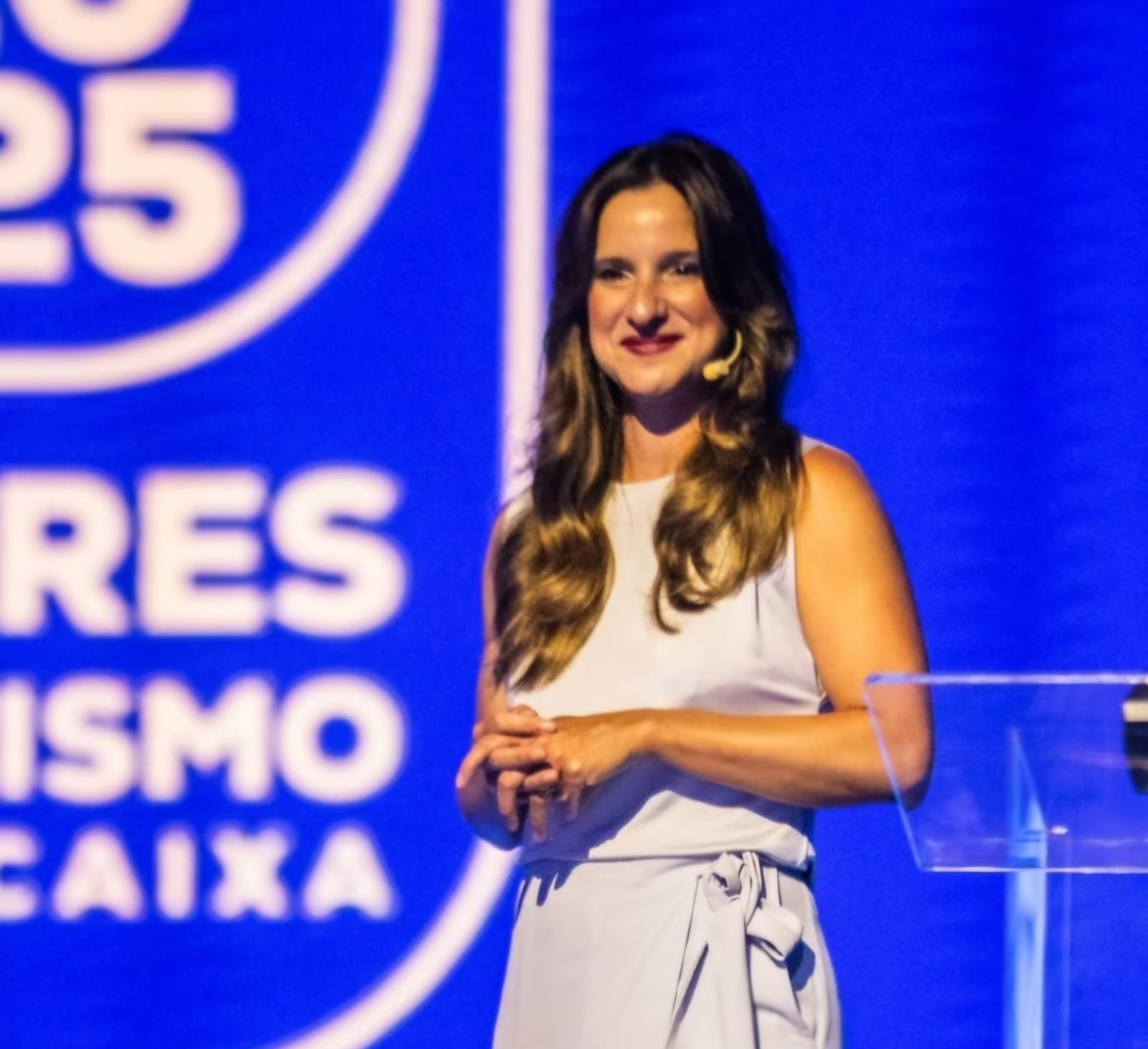 Mulher de cabelos castanhos ondulados, sorrindo, vestindo blusa e saia cinza-claro, com as mãos entrelaçadas à frente. Ela está em um palco com fundo azul e letras brancas que dizem RES ISMO CAIXA